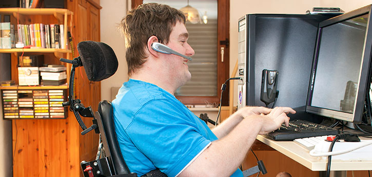 A young mail youth with significant disabilities sits in his wheelchair and uses a Bluetooth headset and keyboard to work on his computer.