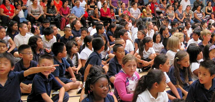 A racially and ethnically diverse group of elementary school students sit in a gymnasium. The group is majority minority.