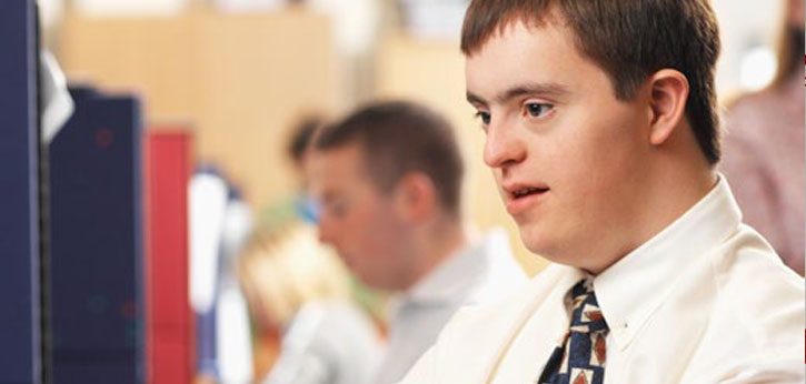 A young man with Down Syndrome in a white button-down shirt and tie works at his desk in an office.