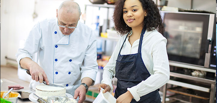 A young woman works in a professional kitchen with a chef.