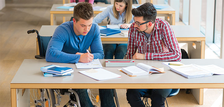 Two older youth, both white males and one in a wheelchair, sit at a table looking over schoolwork together.