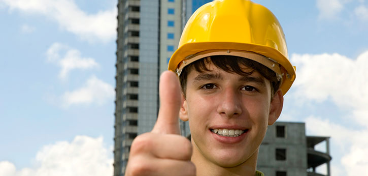 Young man wearing a construction hat gives a thumbs up in front of a tall building.