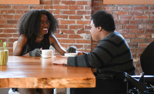image of a young black woman and young black male, dressed in business casual attire, sitting at a coffee shop together, conversing and smiling