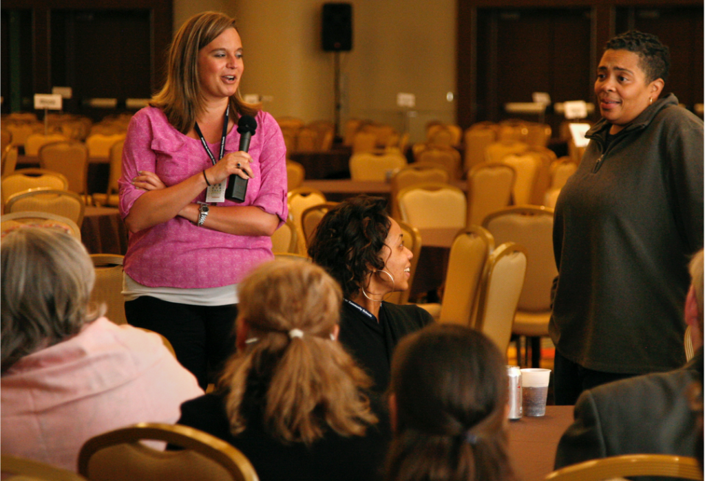 Group of educators around a table. A woman is holding the microphone talking.